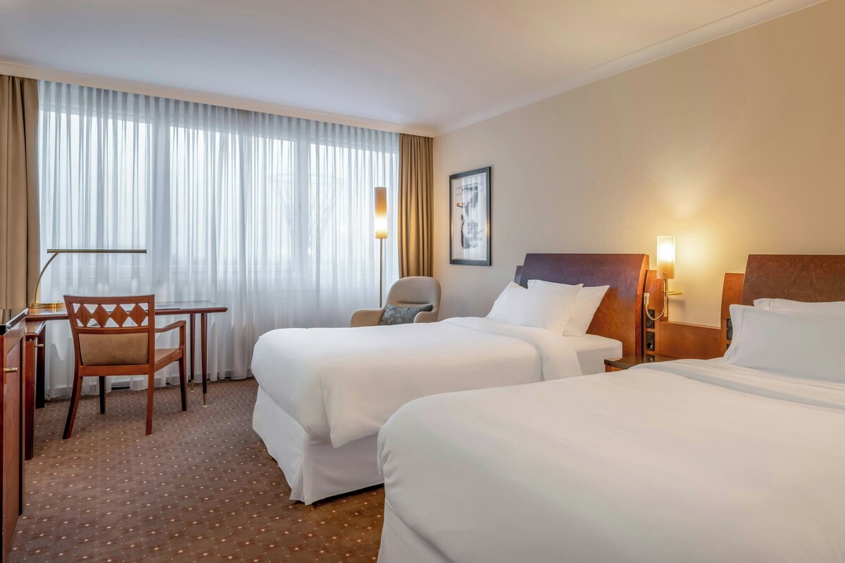 Hotellihuone, jossa on kaksi sänkyä, työpöytä, tuoli ja suuri ikkuna, jossa on verhot.