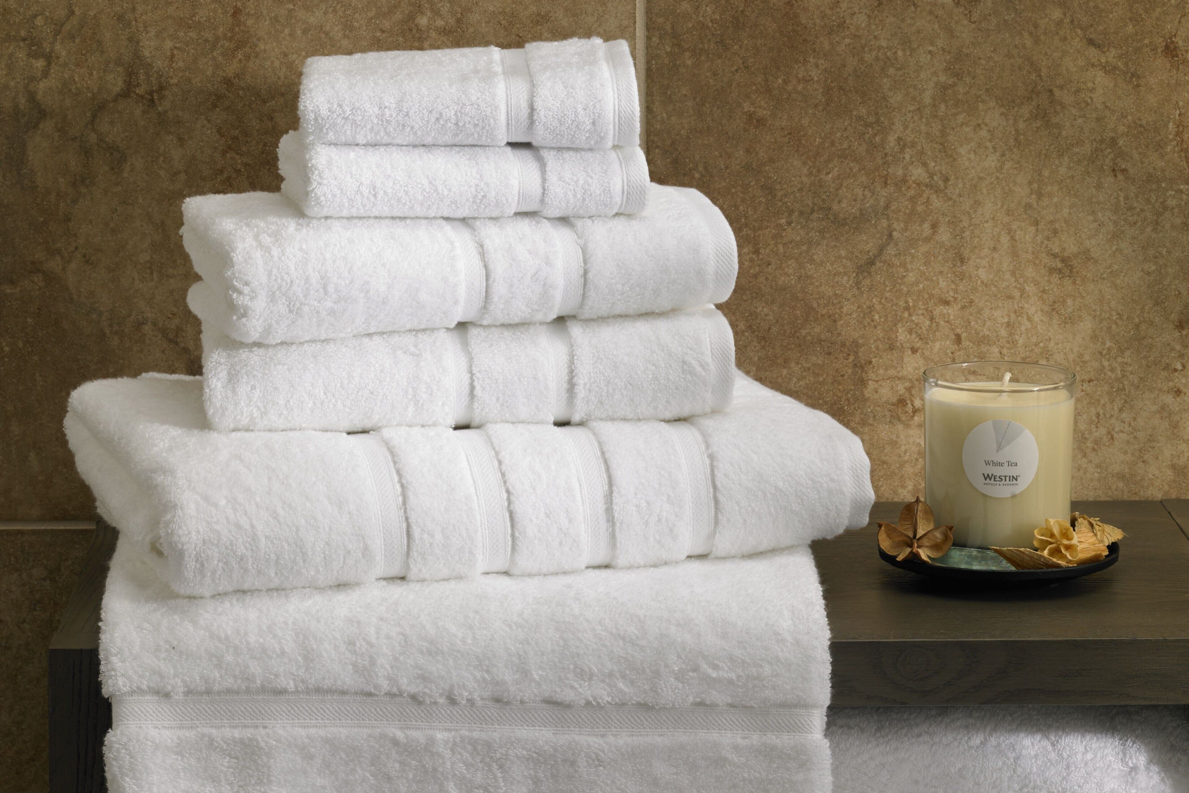 A stack of white towels on a table with a white candle labeled 'White Tea Westin' and dried flowers.