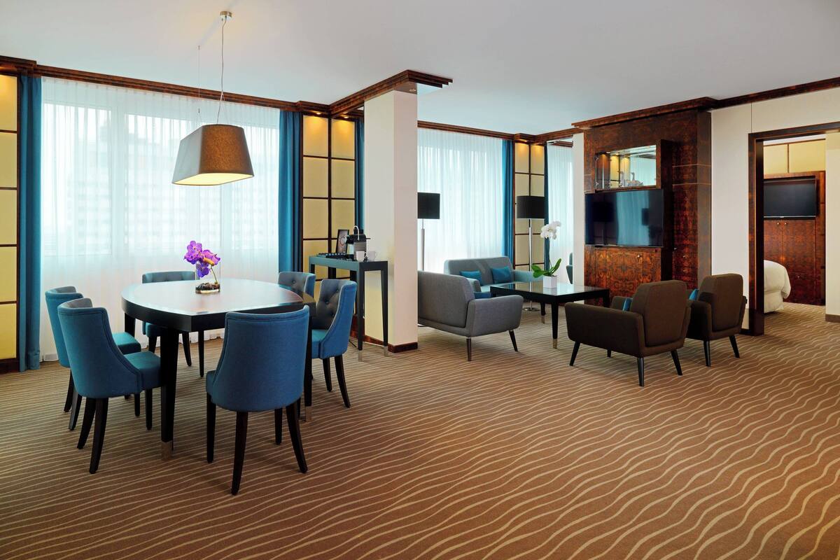 A hotel room with a dining area, a seating area with sofas and chairs, a flat-screen TV, and a large window with curtains.