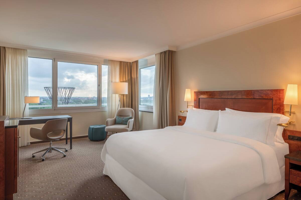 A hotel room with a large bed, a desk, a chair, and a view of the city through large windows.