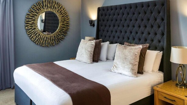 Une chambre d’hôtel avec un grand lit, un miroir rond, une tête de lit sombre et une table de chevet avec une lampe.
