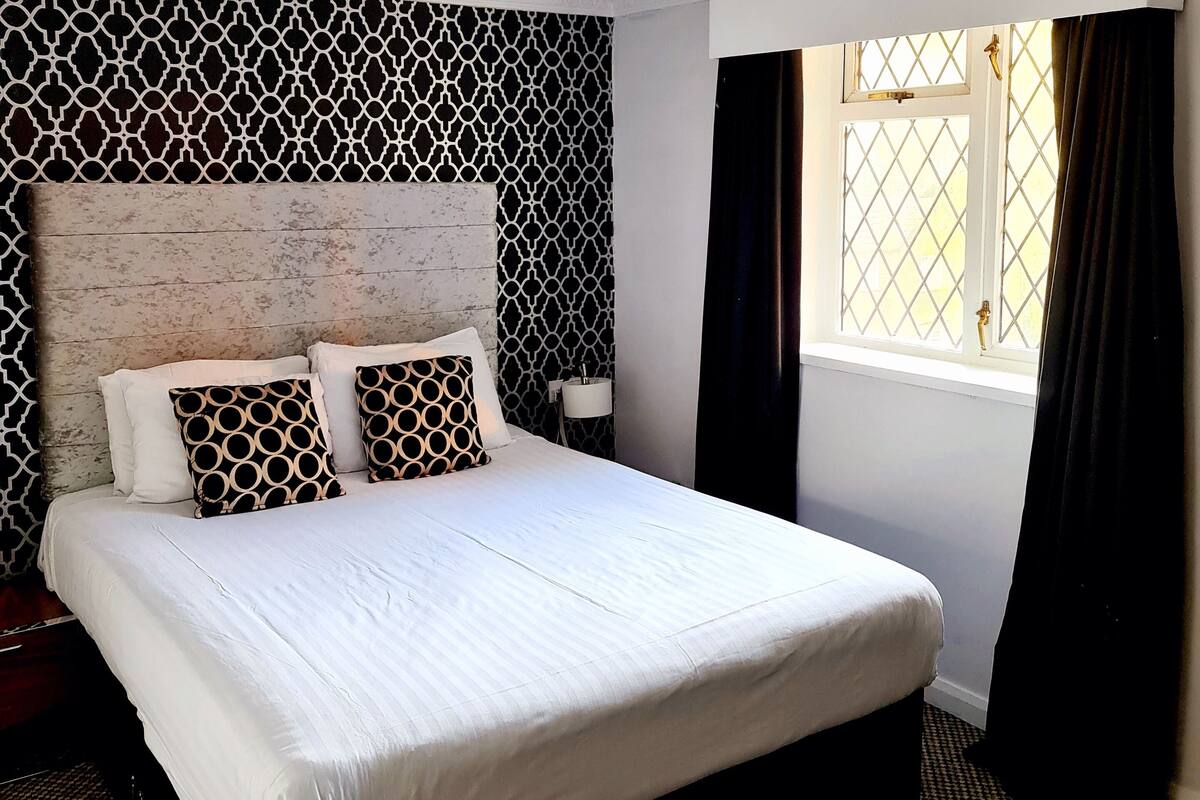 Ein Hotelzimmer mit einem großen Bett, einem gemusterten Kopfteil, einem Fenster mit Vorhängen und einer Wand mit geometrischem Dekor.
