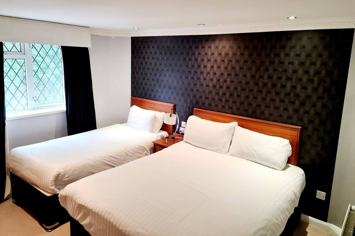 Ein Hotelzimmer mit zwei Einzelbetten, einem dunkel gesteppten Kopfteil, einem Fenster mit Vorhängen und einem Nachttisch mit Lampe.