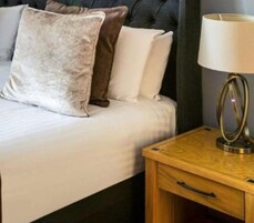 Una cama con almohadas blancas y marrones, una mesita de noche con una lámpara y una mesita auxiliar de madera.