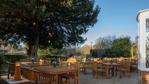 Un espace extérieur aménagé en terrasse, avec des tables et des chaises en bois, entouré d’arbres et d’un mur en briques.