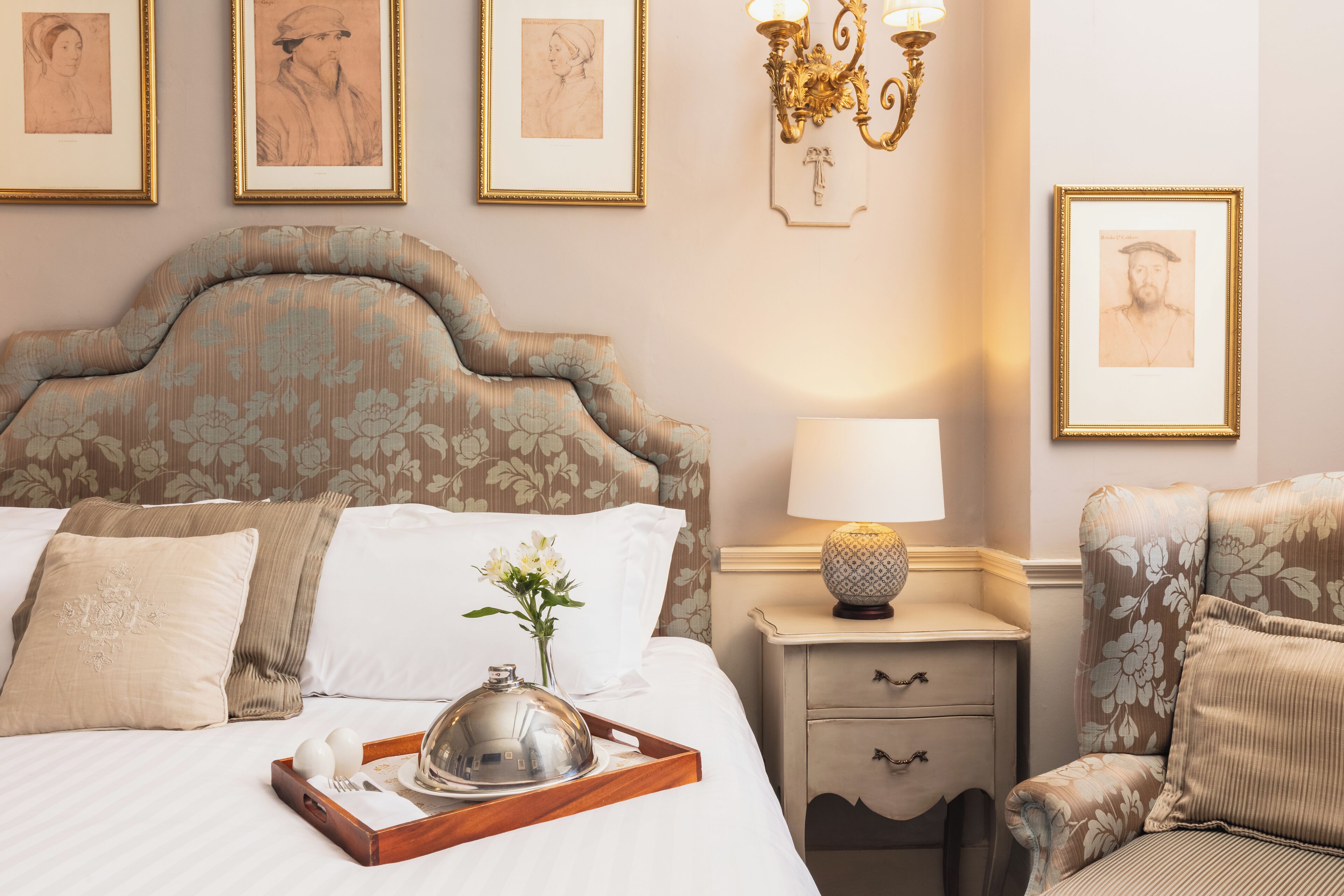 A neatly made bed with a tray, pillows, and a vase of flowers. There are framed pictures on the wall and a lamp on the nightstand.