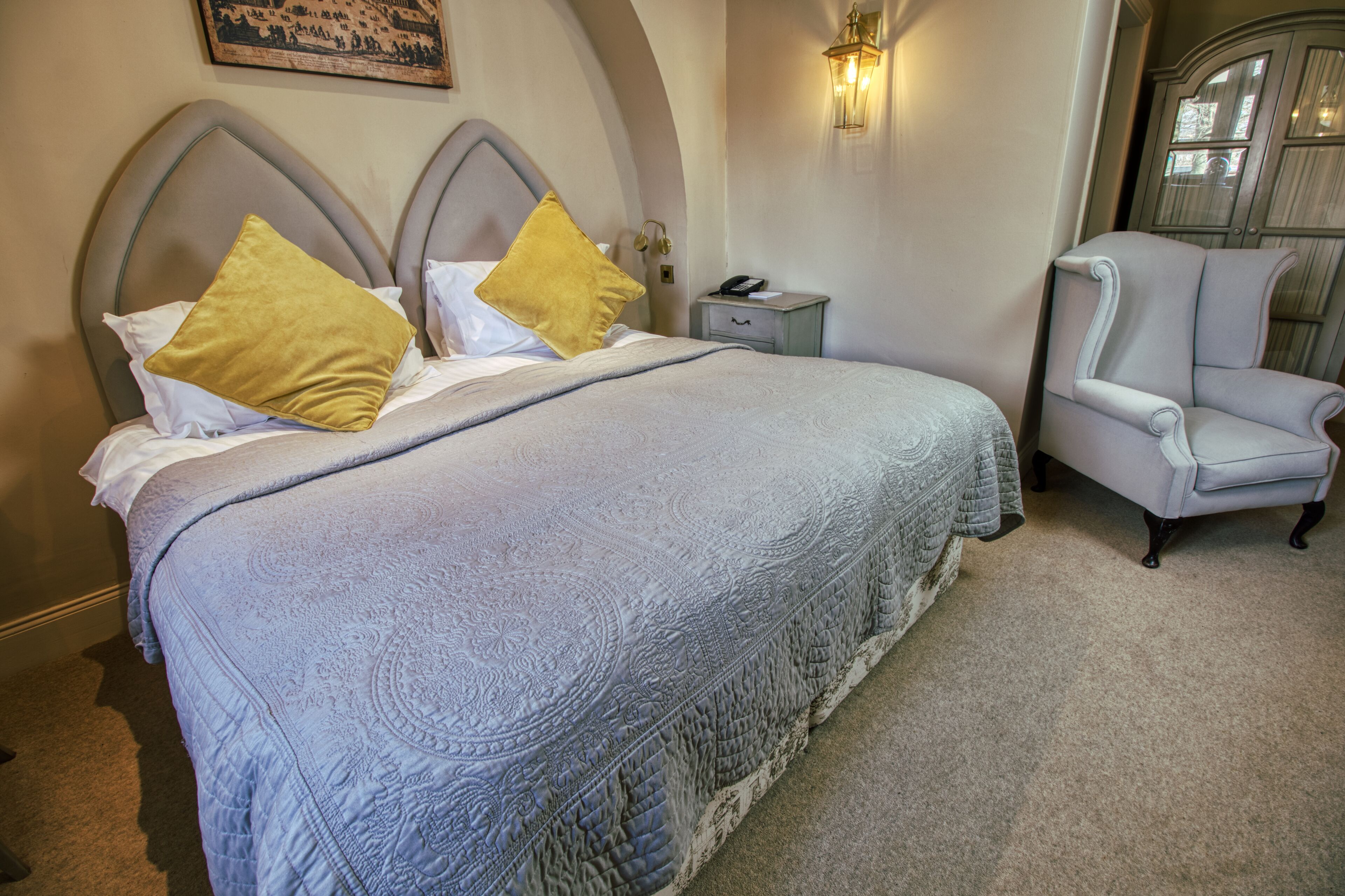 A neatly made bed with a patterned bedspread and two gold pillows, a small bedside table with a lamp, and a classic armchair.