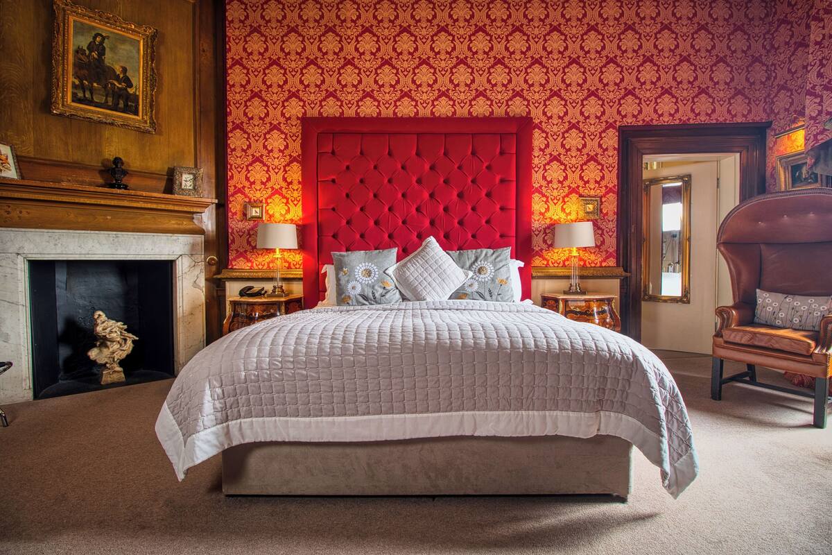 A bedroom with a large bed, a red headboard, two bedside tables with lamps, a fireplace, and a chair.