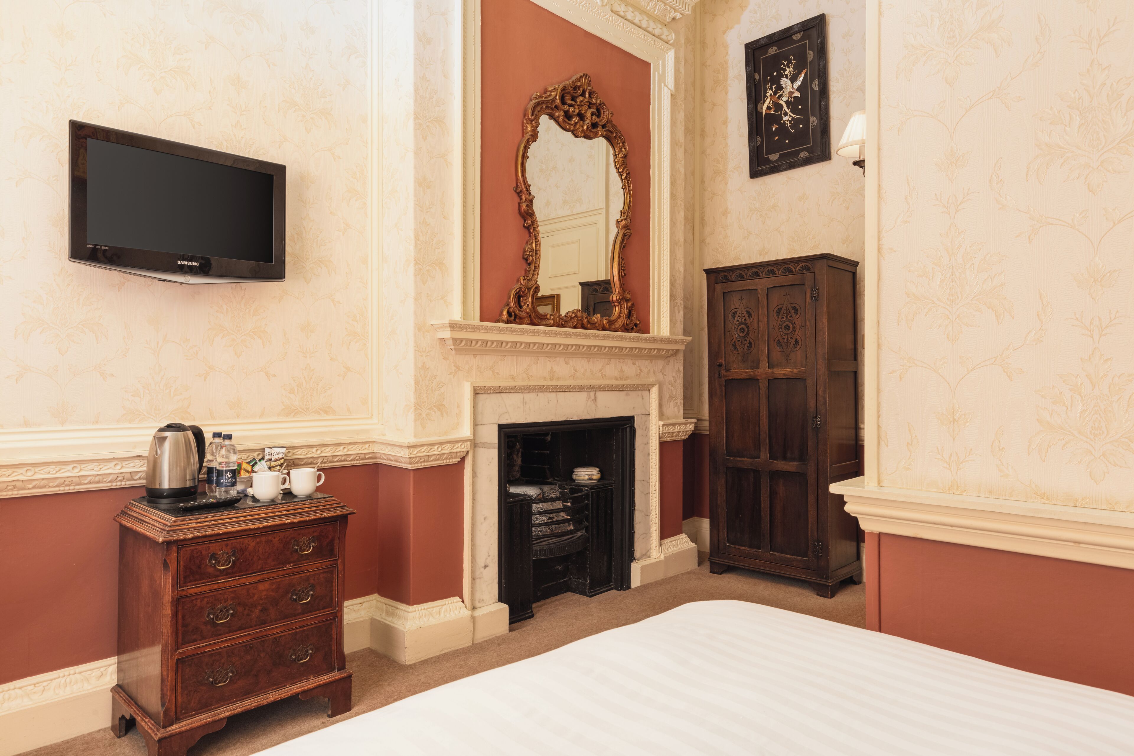 A hotel room with a bed, a flat-screen TV, a fireplace, a wooden cabinet, and a large mirror.
