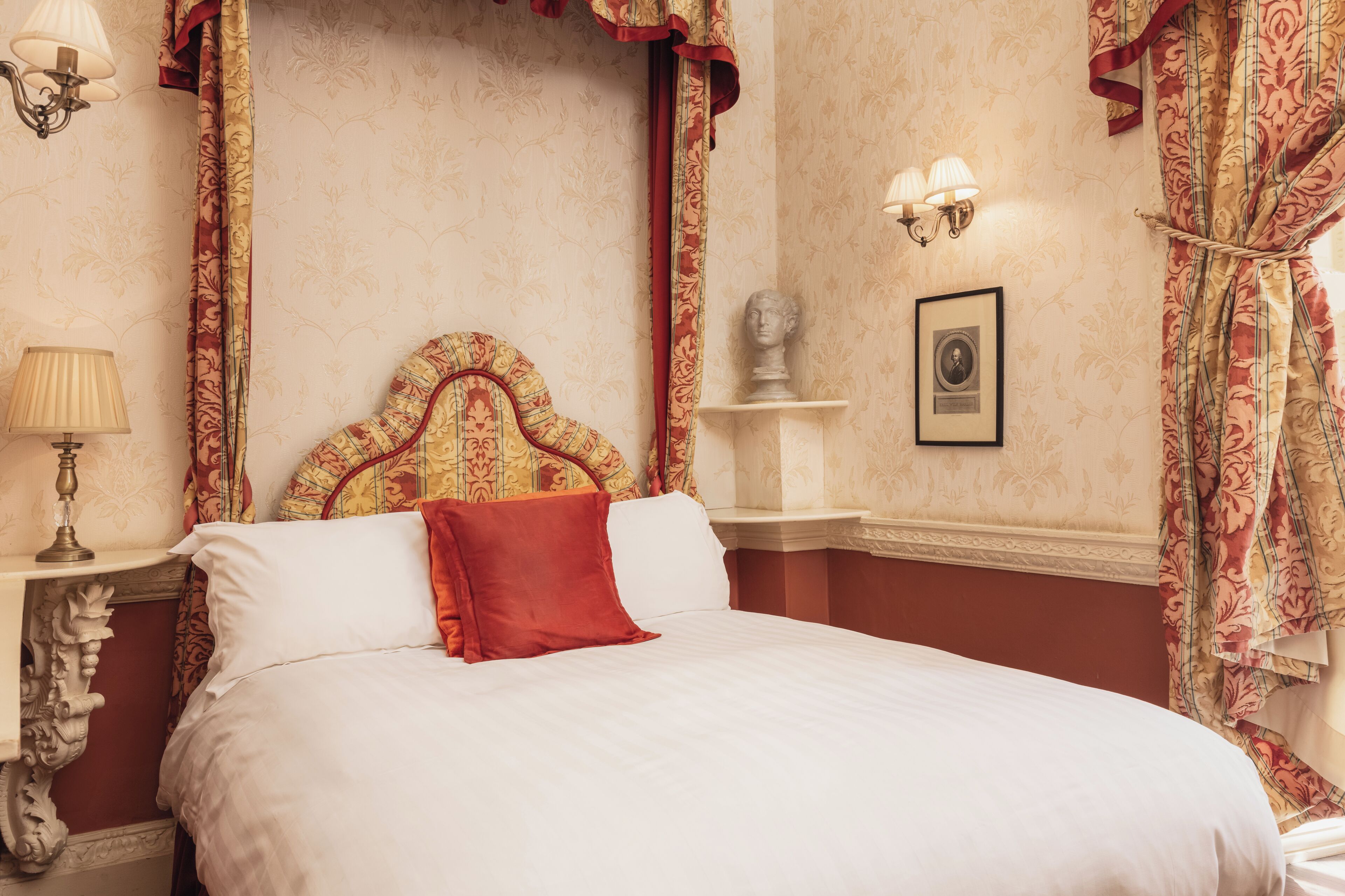 A classic hotel room with a large bed, ornate curtains, a bust on a shelf, and wall sconces.