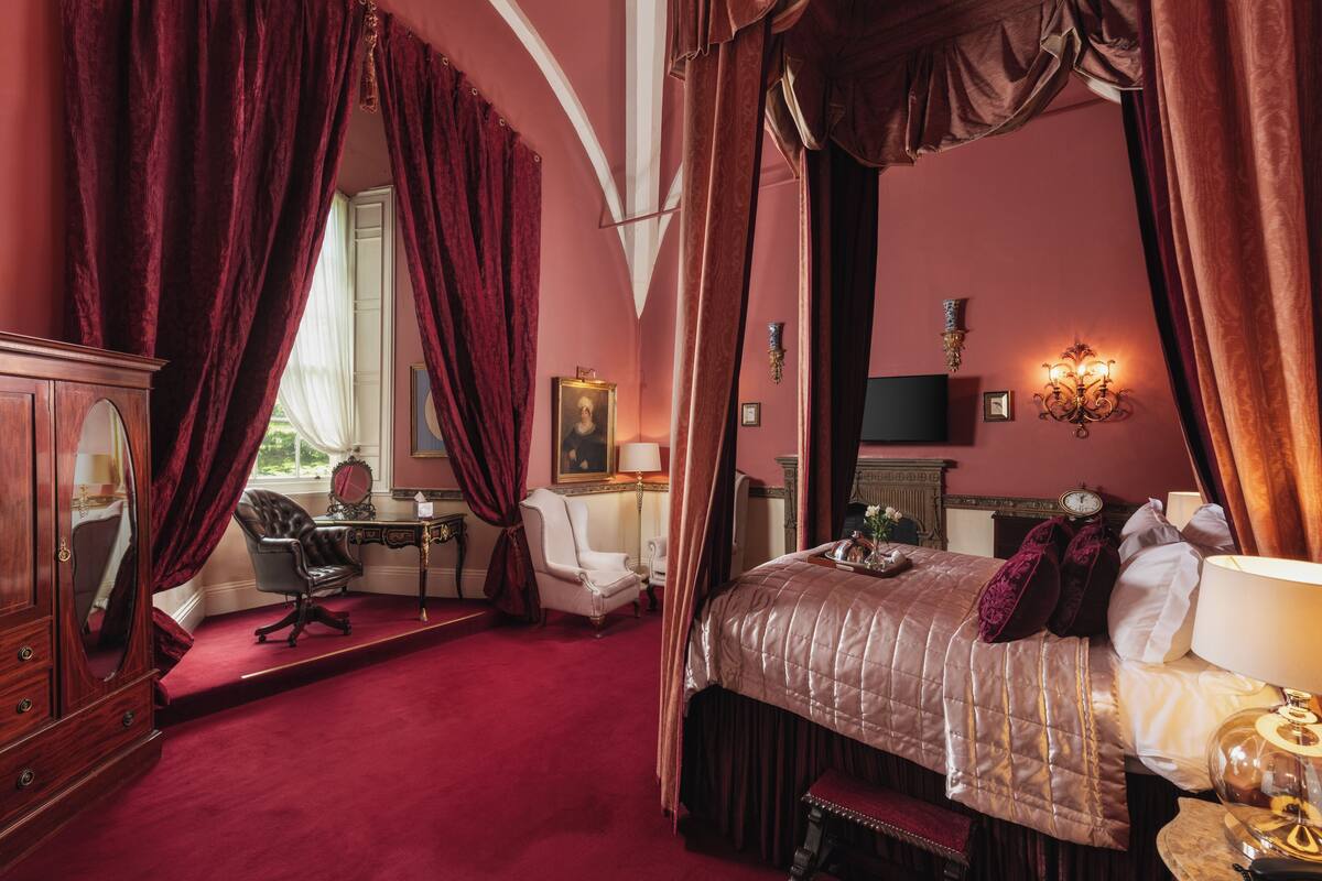 A bedroom with a four-poster bed, a desk with a chair, a mirror, and a fireplace.
