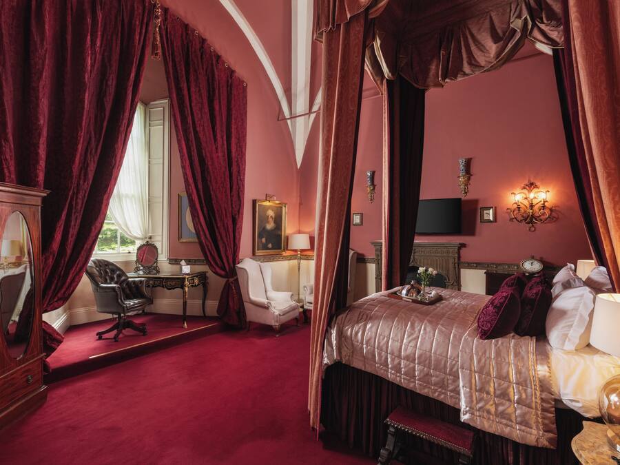 A bedroom with a four-poster bed, a desk with a chair, a mirror, and a fireplace.