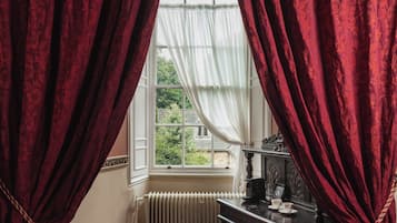 Una habitación con cortinas rojas, una ventana con vistas a la vegetación, un radiador y una mesa con tetera.