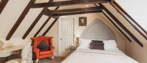 A bedroom with a sloped ceiling, a bed, a desk, a chair, and a small table with a lamp.