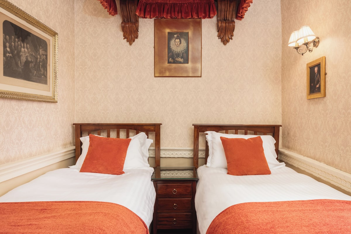 A hotel room with two beds, a wooden nightstand, a framed picture above the beds, and a wall-mounted light fixture.