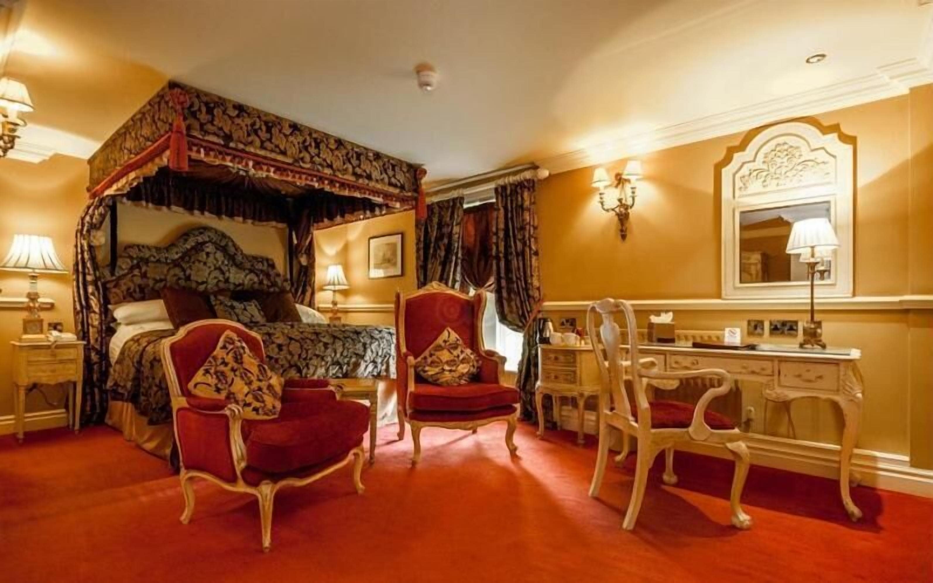 Ein traditionelles Himmelbett mit Baldachin, zwei rote Polsterstühle, ein weißer, verziierter Schreibtisch und ein Spiegel an der Wand.