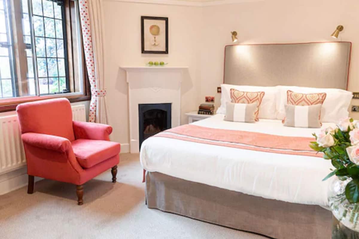 A bedroom with a large bed, a red armchair, a fireplace, and a window with curtains.