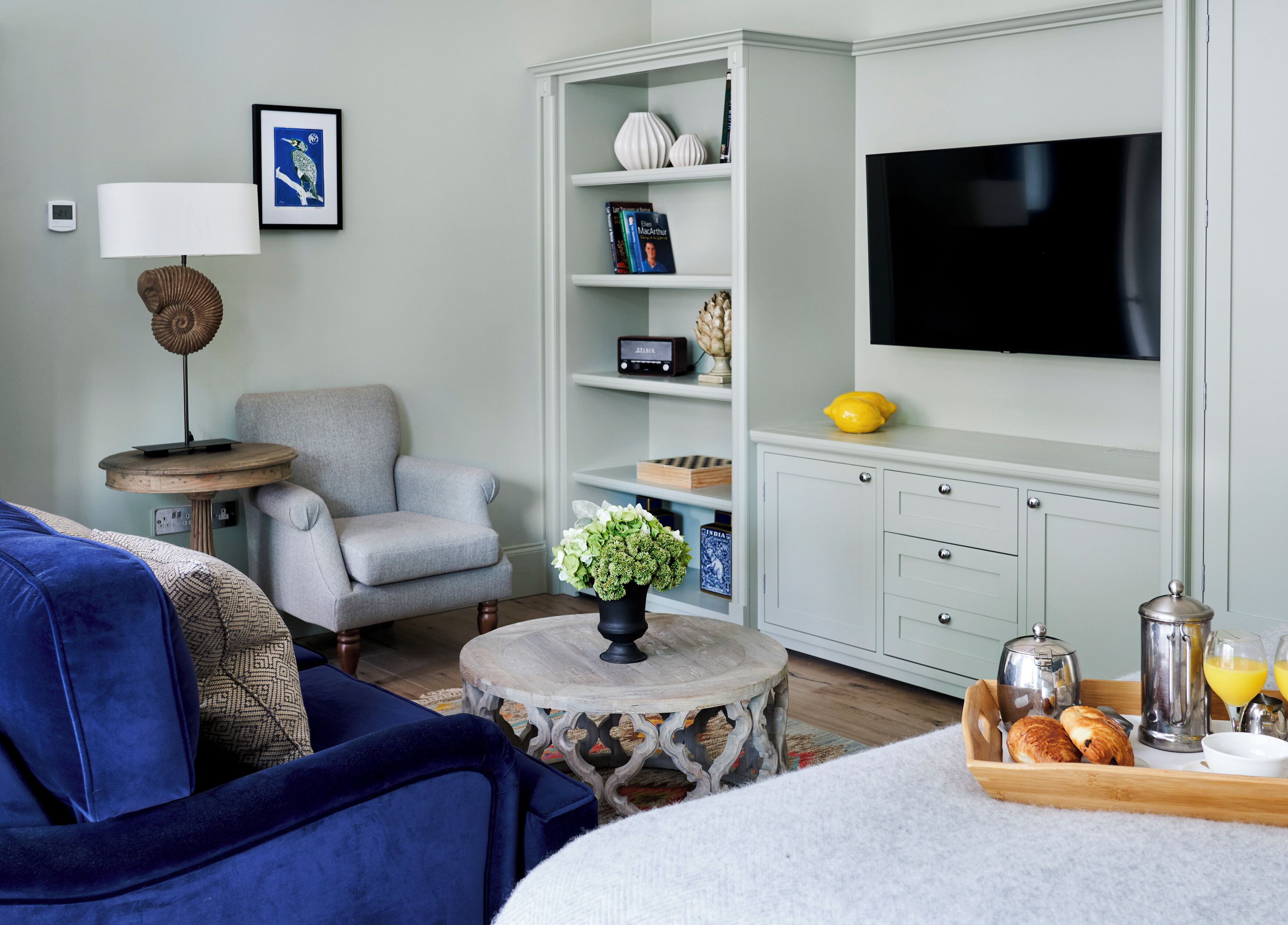 A modern living room with a grey armchair, a round coffee table with a vase, a bookshelf, a flat-screen TV, and a breakfast tray with croissants and juice.