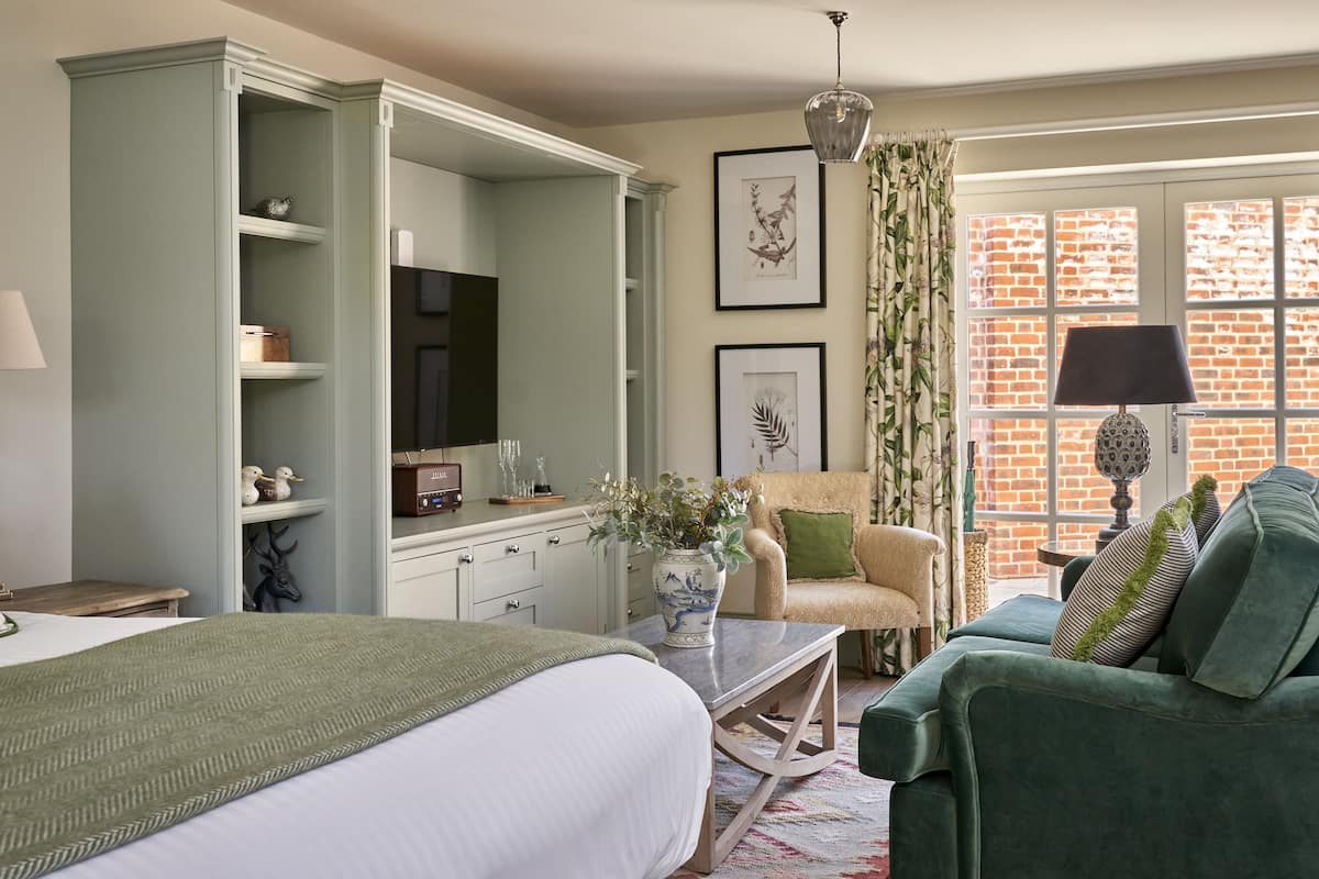 A bedroom with a bed, a dresser, a vanity, a green armchair, a green sofa, and a window with a brick wall view.