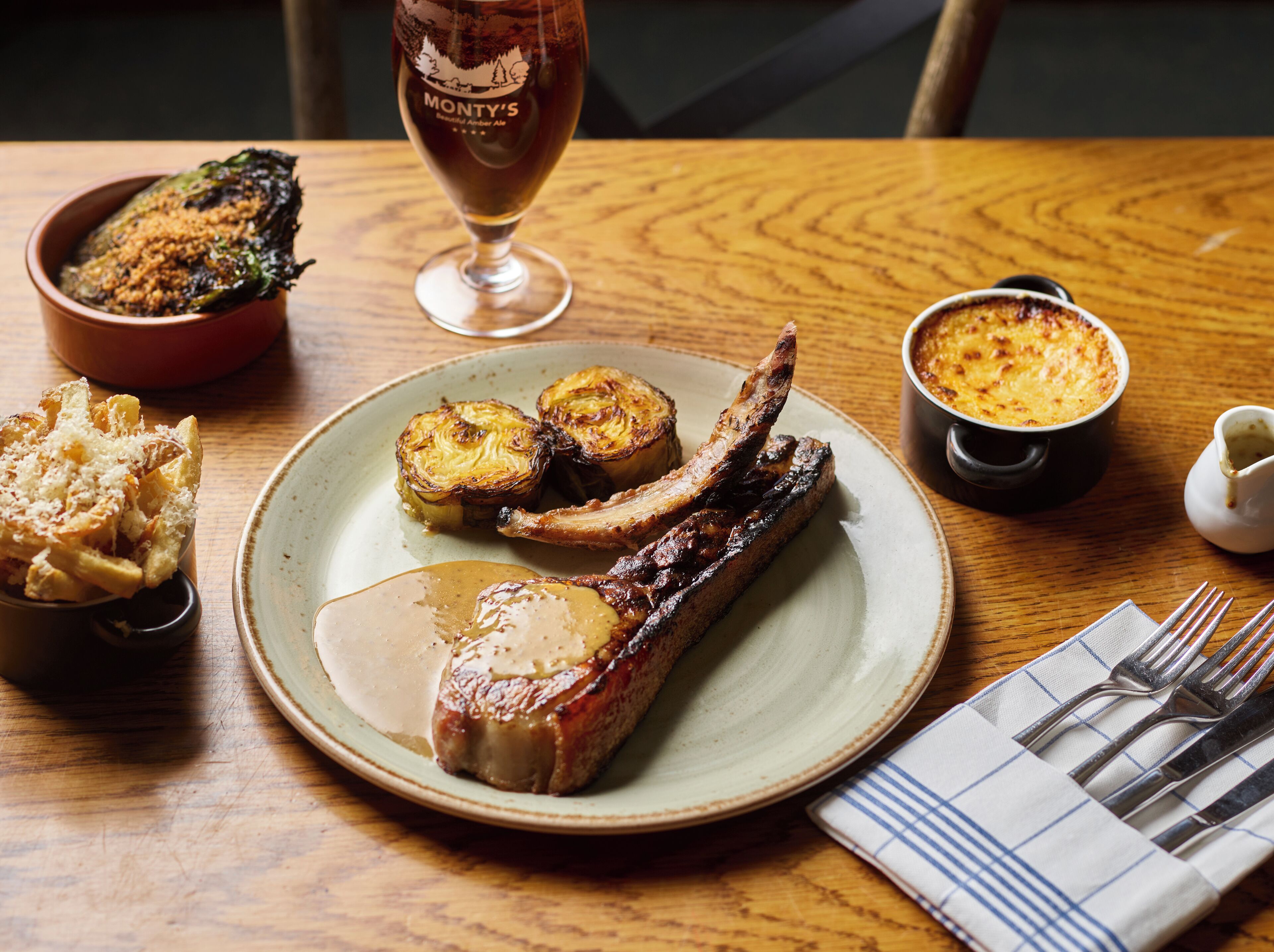A dining table with a plate of roasted meat, a bowl of fries, a glass of beer, and a small pot of sauce.