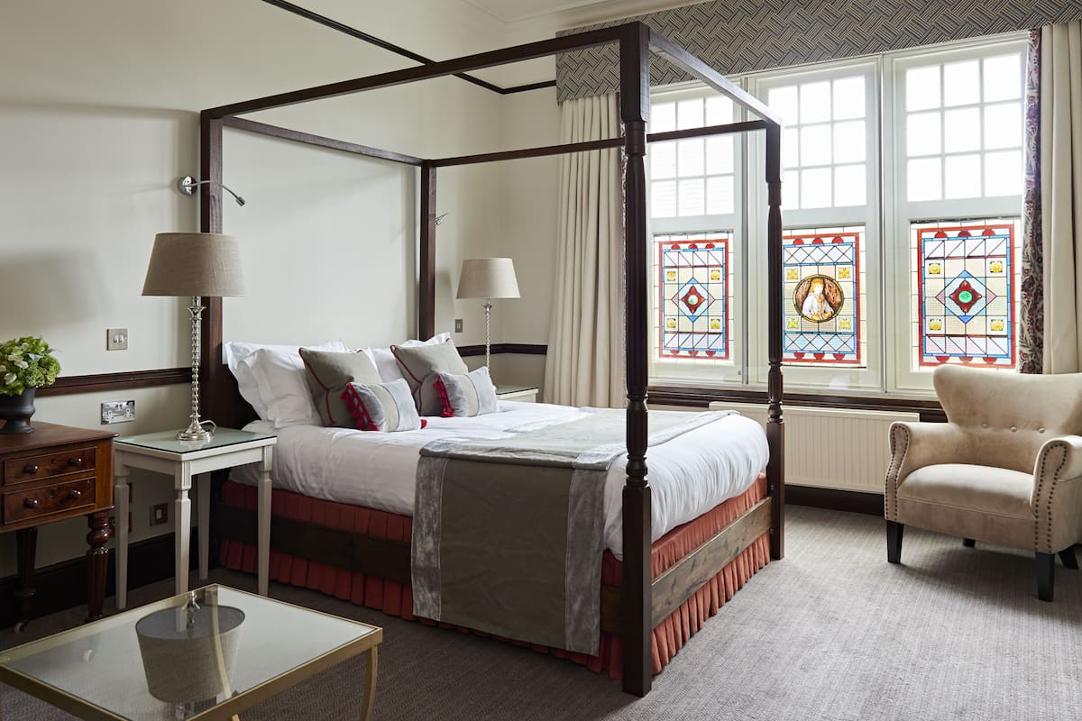 A four-poster bed with a canopy, two bedside tables, a chair, and a view of stained glass windows.
