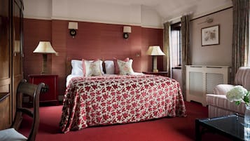 A hotel room with a large bed, two bedside tables with lamps, a wooden chair, a small table with a vase, and a radiator.