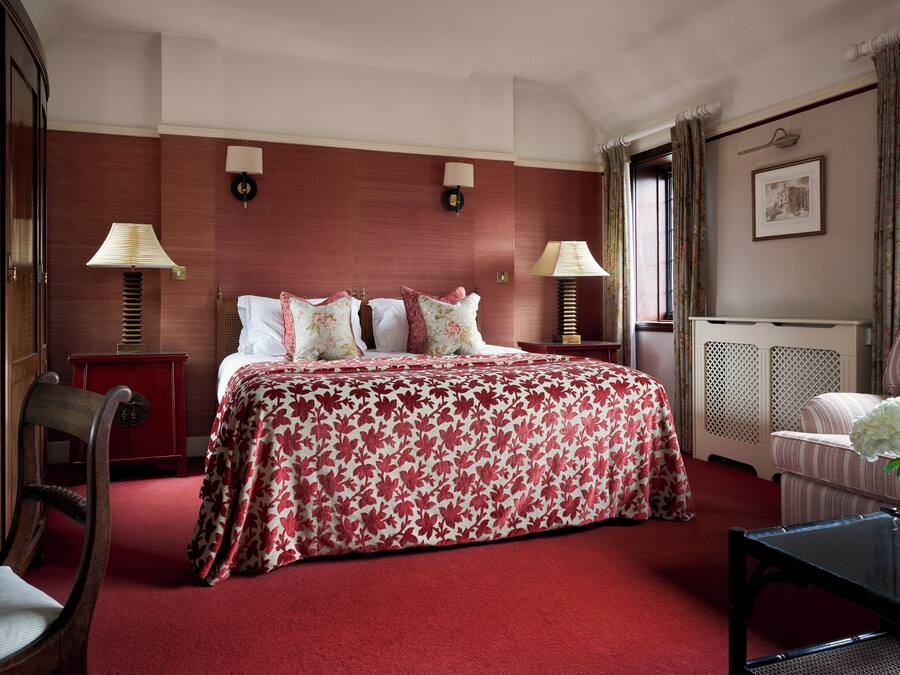 A hotel room with a large bed, two bedside tables with lamps, a wooden chair, a small table with a vase, and a radiator.