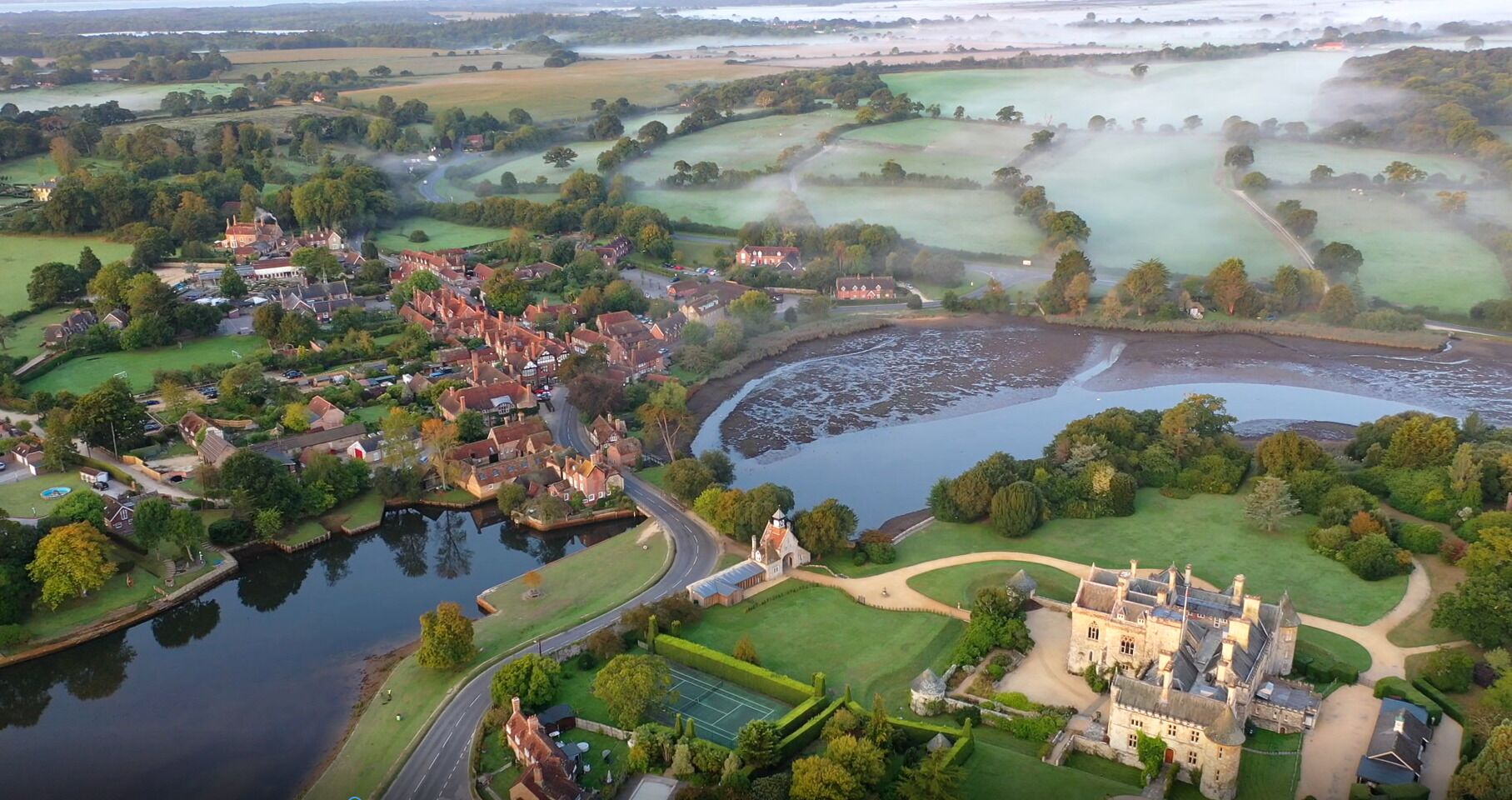 An aerial view of a riverside town with a prominent castle, residential areas, and green fields.