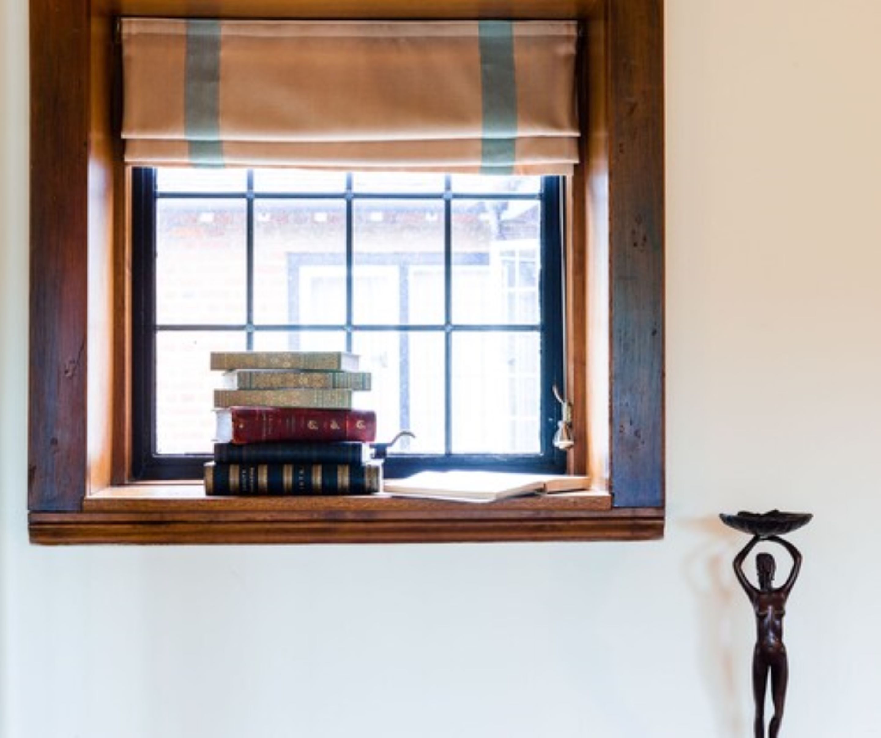A window with a view of a building, a stack of books on a ledge, and a sculpture on the wall.