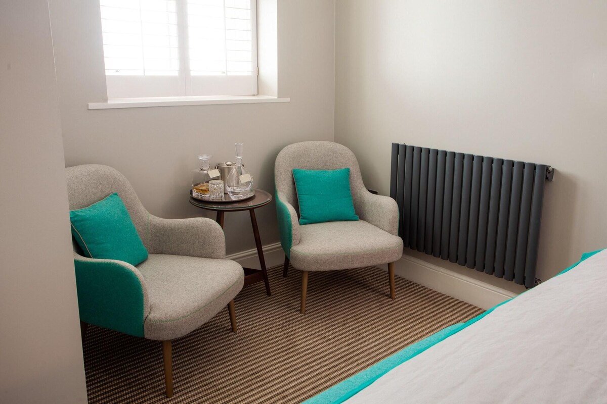 A small room with two grey armchairs, each with a teal cushion, a small round table with a tray of items, and a radiator against the wall.