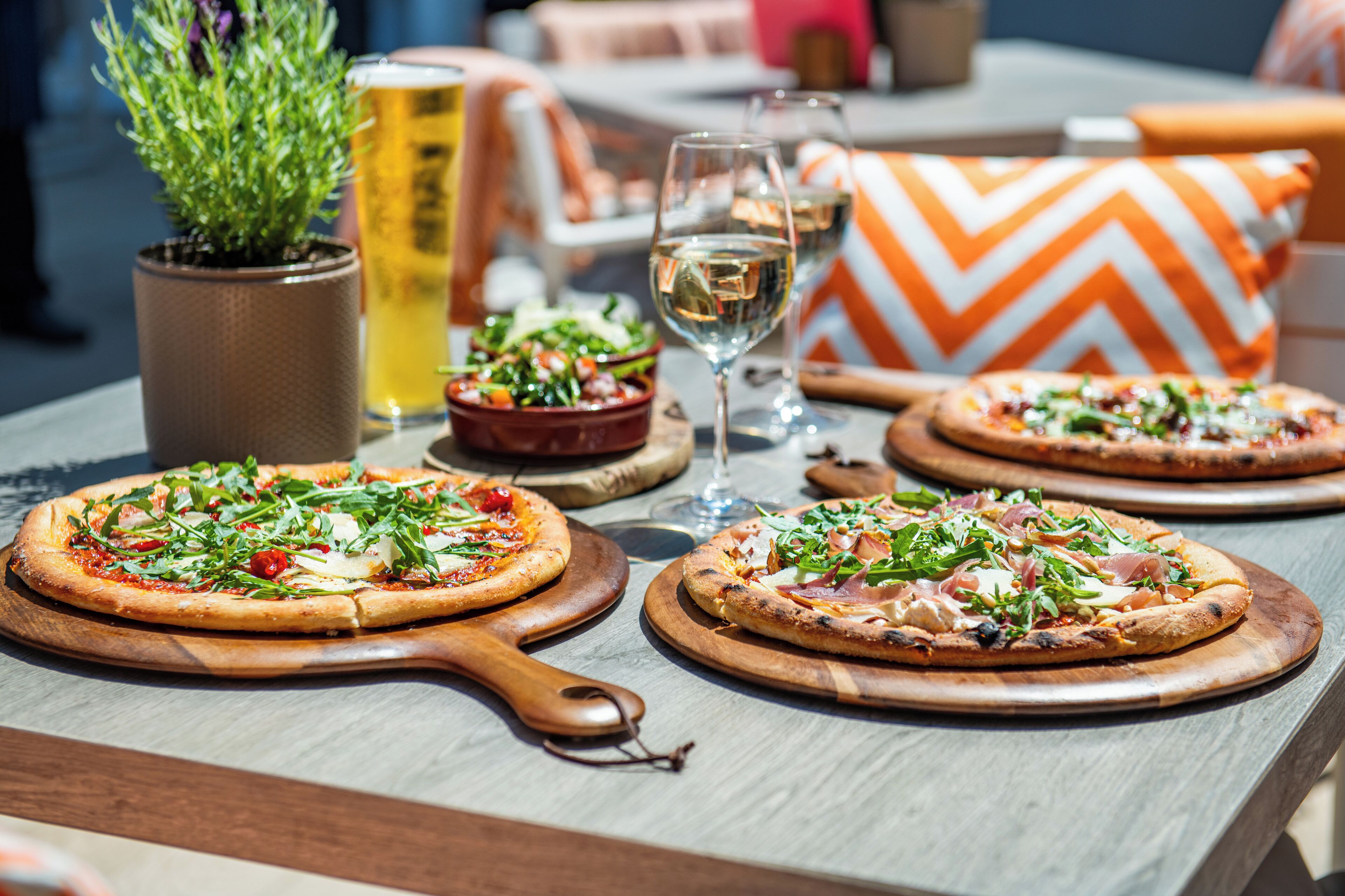 Drie pizza's op houten planken, met een potplant en een glas bier op een tafel.