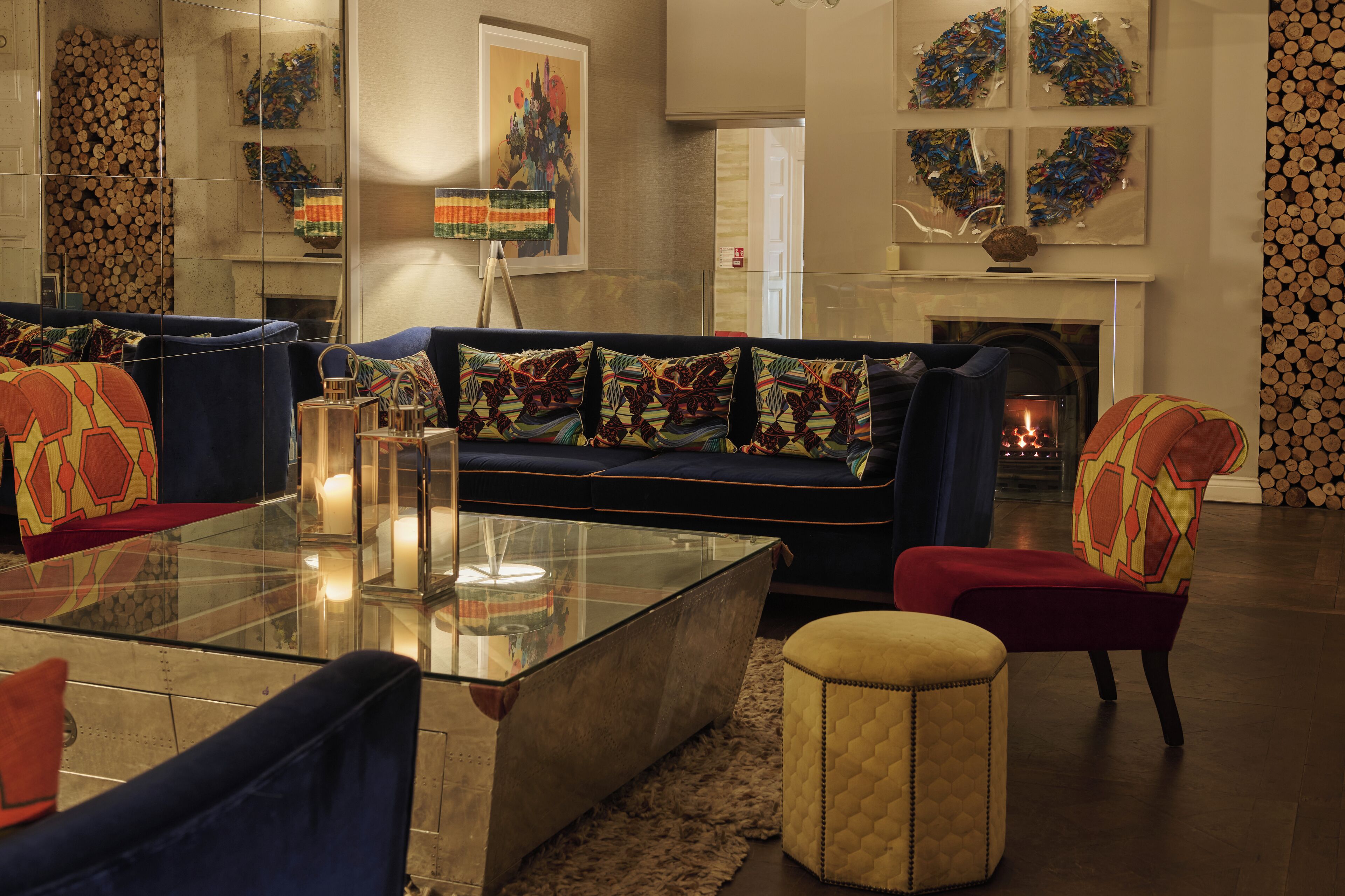 A modern living room with a dark blue sofa, colorful patterned cushions, a glass coffee table, and a small ottoman.