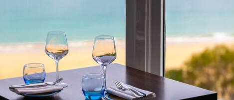 A table with a glass of water, a wine glass, and a folded napkin with cutlery, set against a window with a view of the beach.