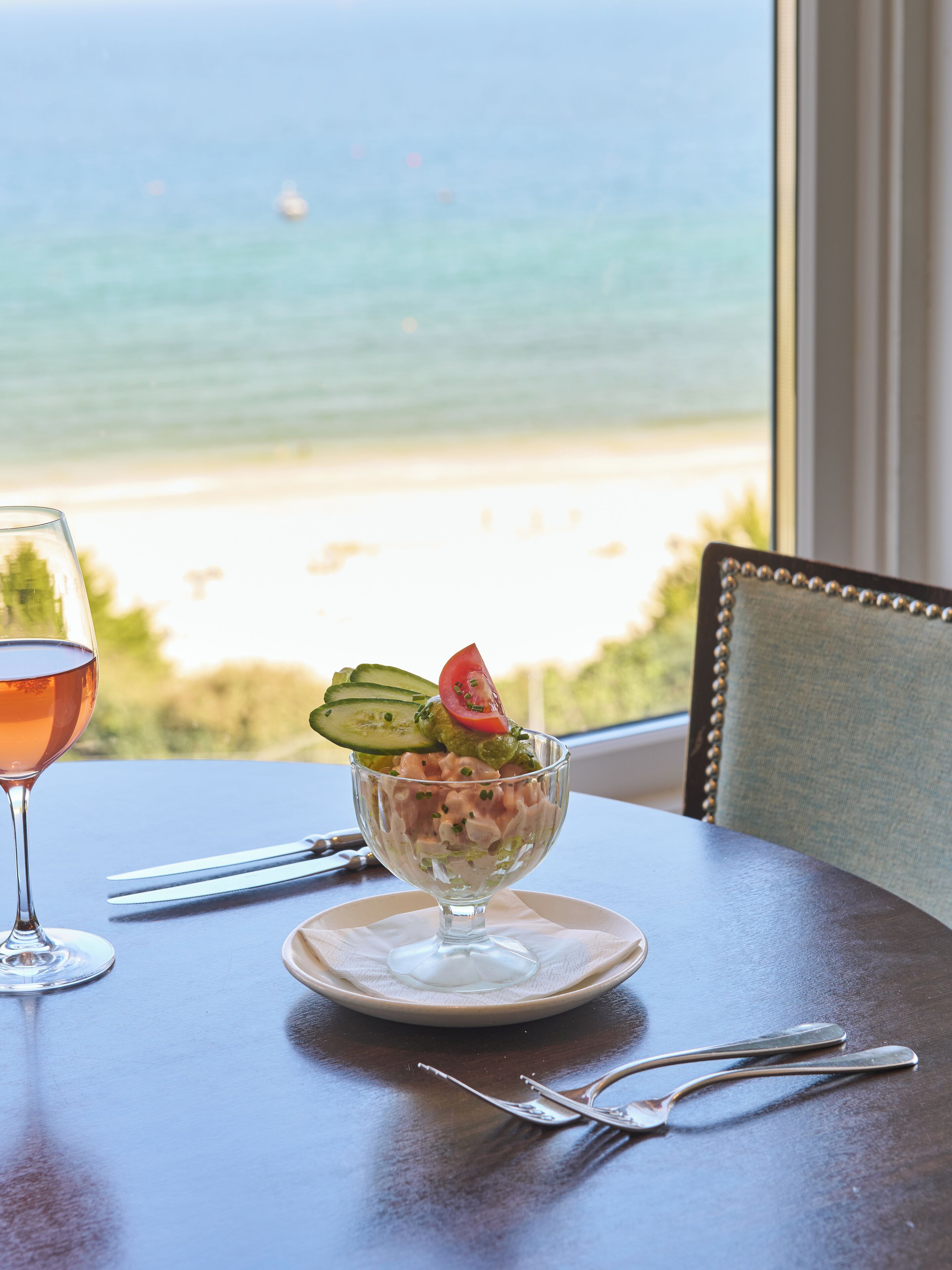 Una mesa con un bol de postre, una copa de vino y cubiertos. Una ventana muestra una vista a la playa.