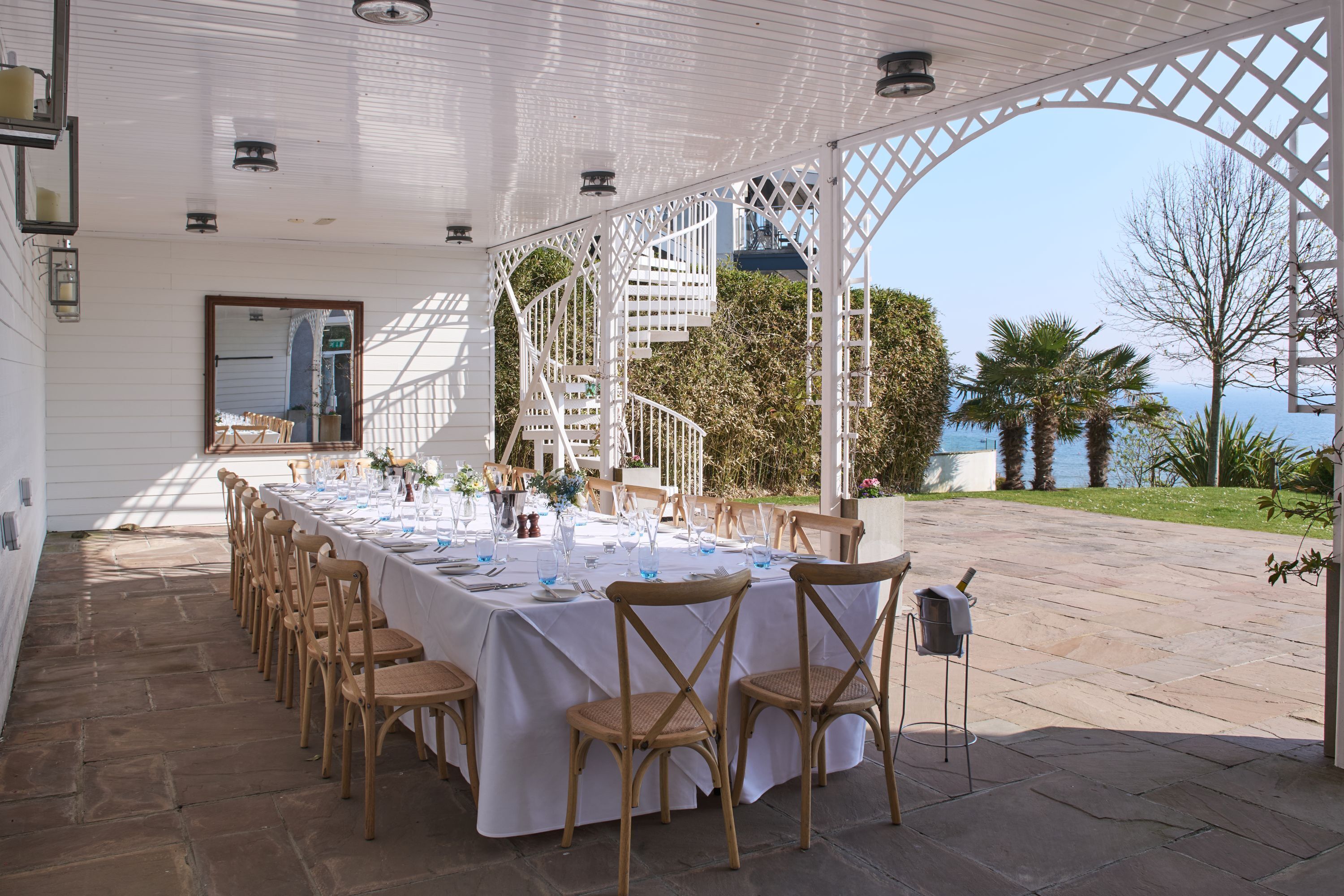 An outdoor dining area with white tables and chairs, a staircase, and a view of the sea.