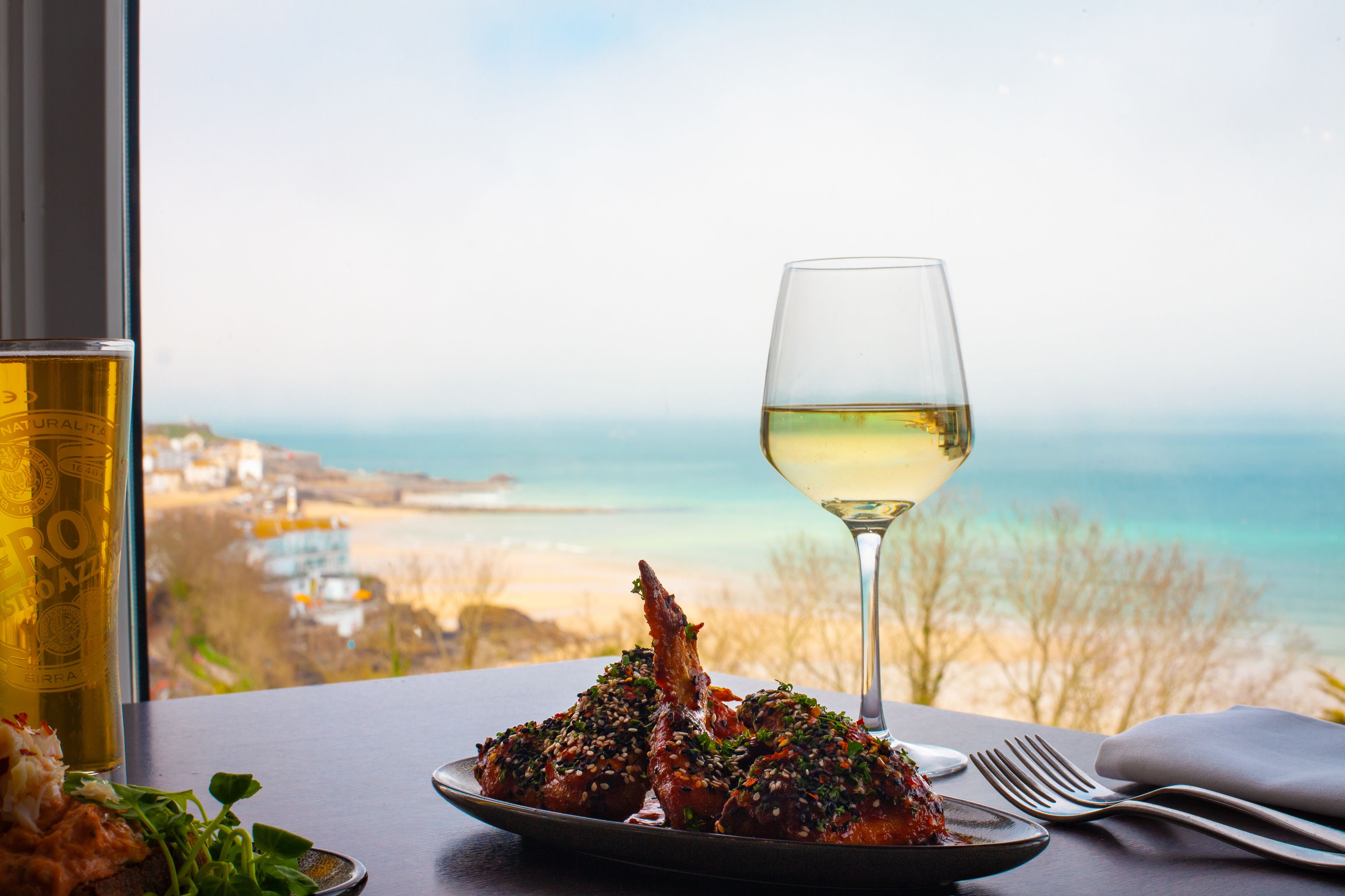 A table with a plate of food, a glass of white wine, and a view of the beach.