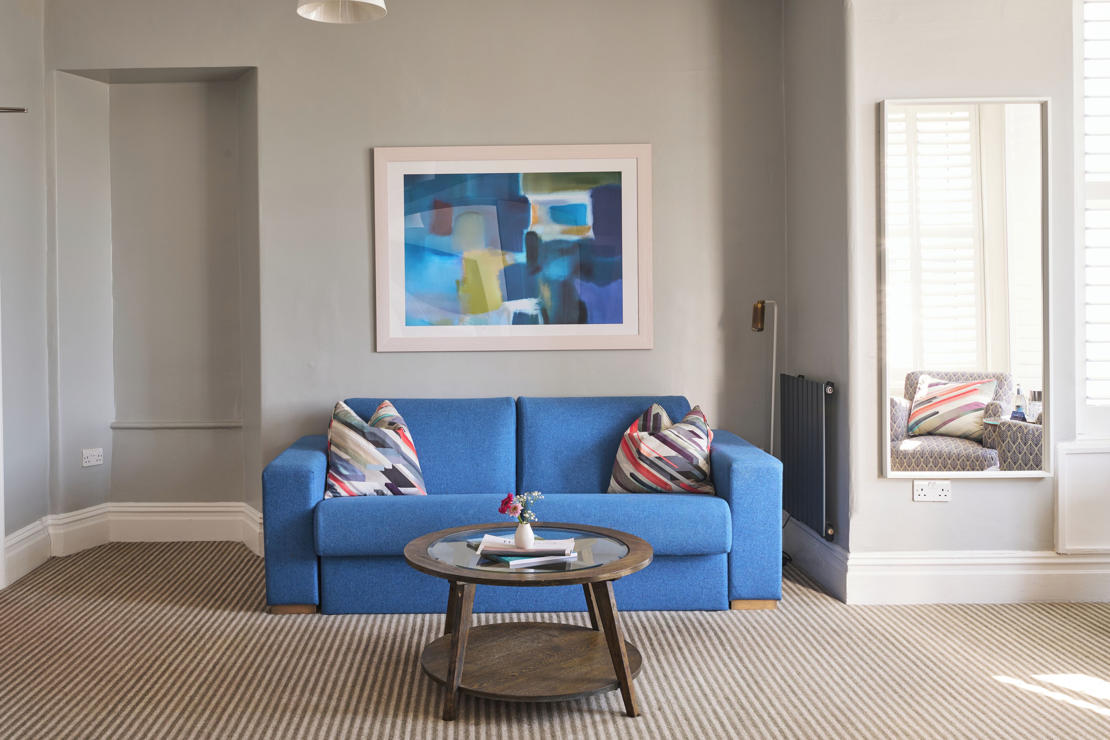 A living room with a blue sofa, a round coffee table, a framed abstract painting, and a window with blinds.