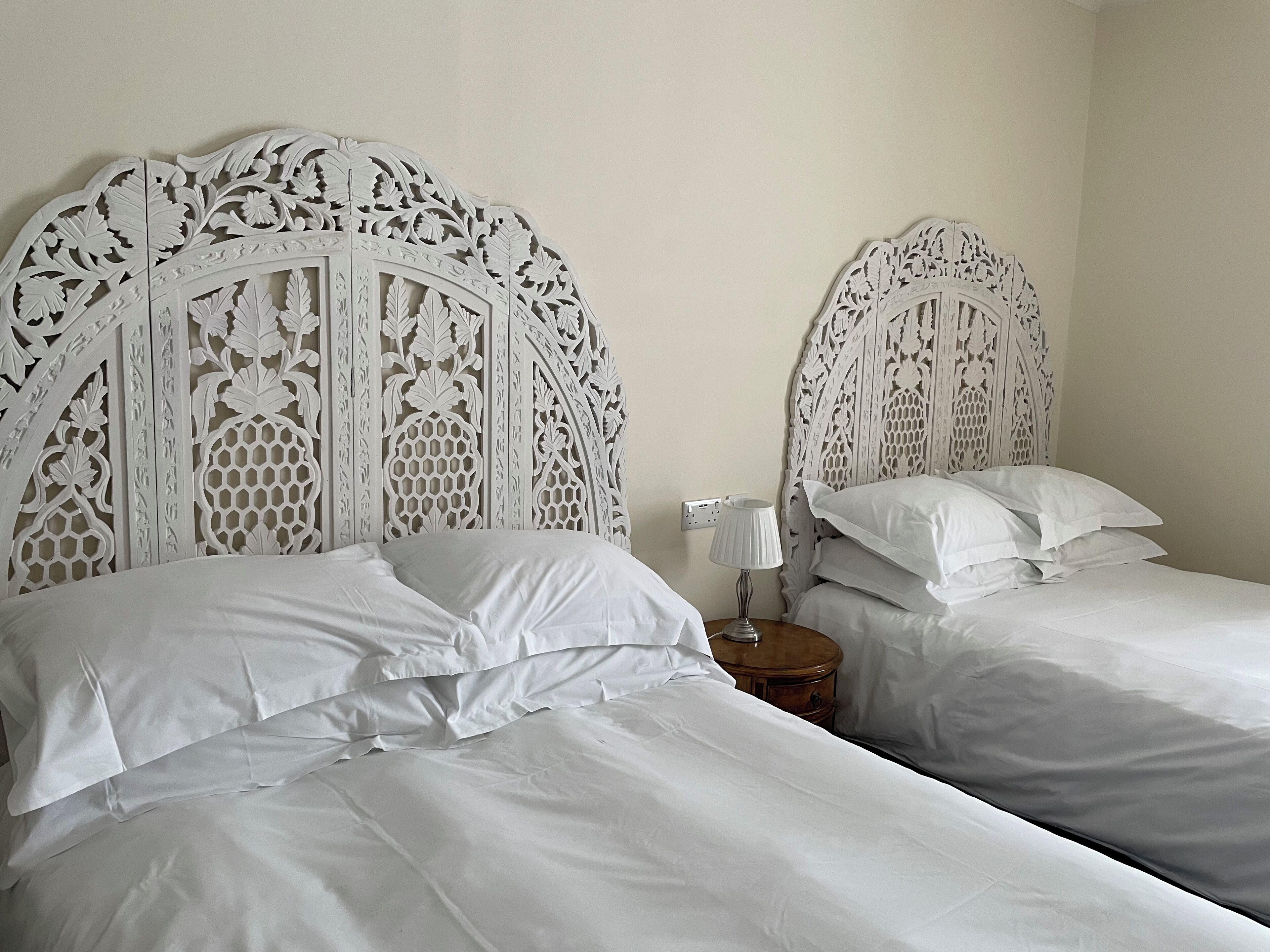 Two beds with white linens and ornate headboards, a bedside table with a lamp, and a power outlet on the wall.
