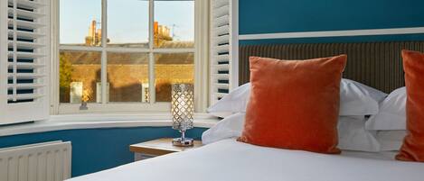 A bedroom with a large window, white shutters, a bedside table with a lamp, and orange pillows.