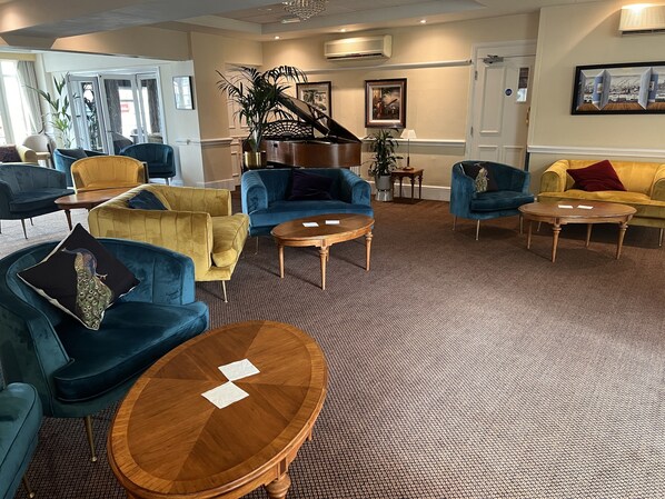 A spacious lounge area with multiple seating options including sofas and armchairs, a grand piano, and a carpeted floor.