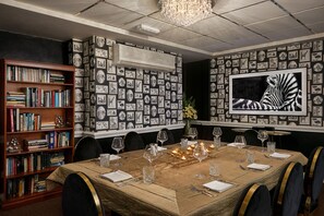 A dining area with a long table set for six, surrounded by chairs. A bookshelf filled with books is to the left. The wall features a large framed zebra portrait and a patterned wallpaper with black and white images. - The Royal Adelaide Hotel (Windsor)