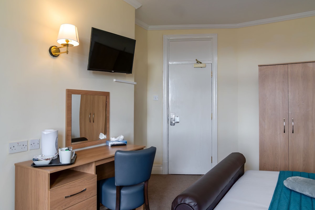 Habitación de hotel con un escritorio de madera, una silla, una cama, un televisor montado en la pared y un armario.