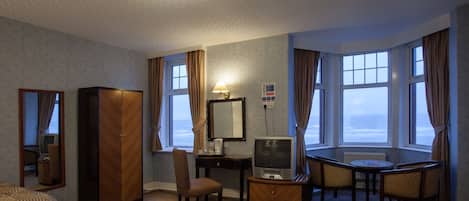 Hotellihuone, jossa on sänky, työpöytä tietokoneella, tuoli, pieni pöytä ja suuret ikkunat, joista avautuu näkymä merelle.