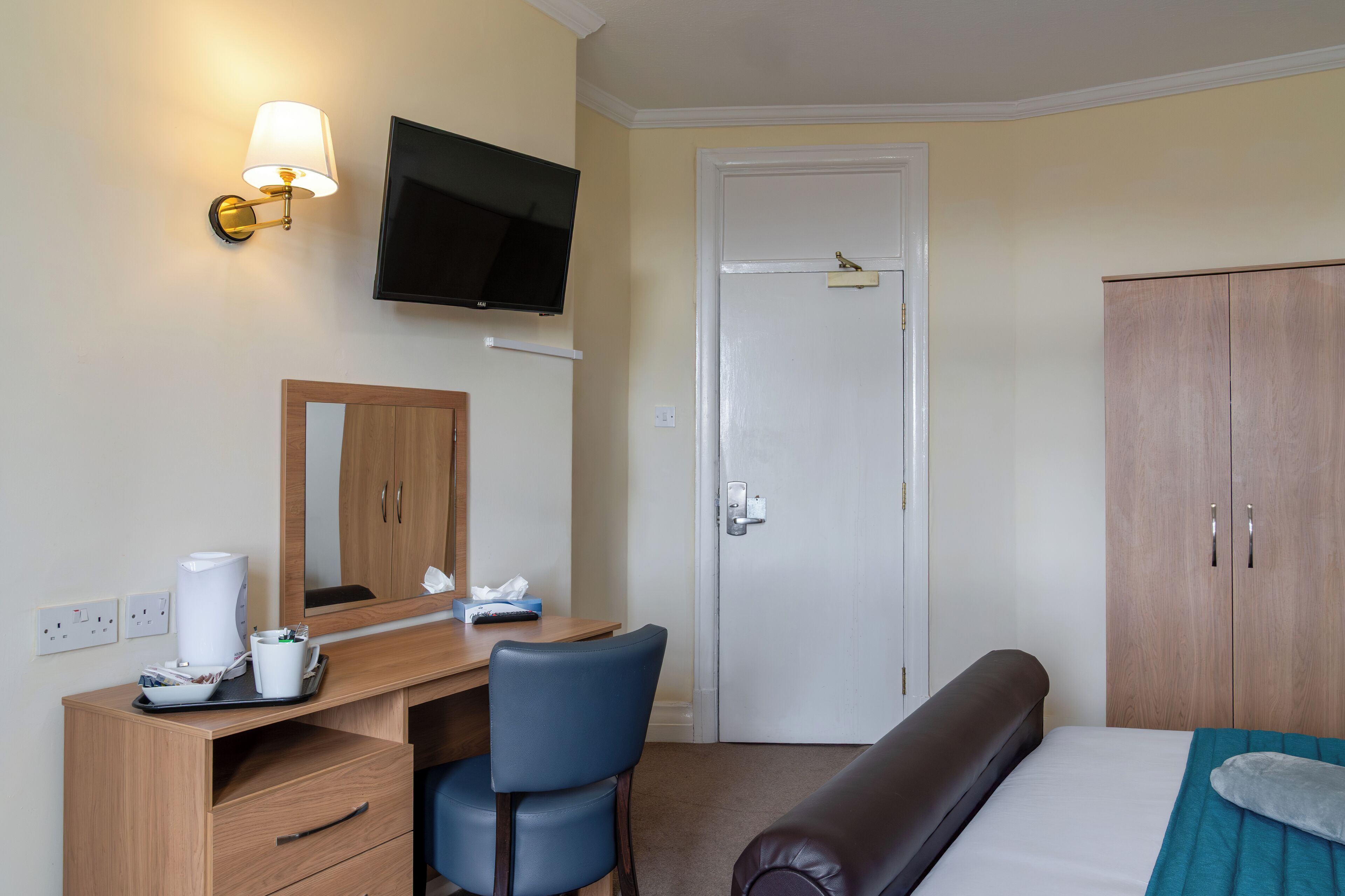 Habitación de hotel con escritorio de madera, una silla, cama con manta color turquesa, televisor montado en la pared y un armario.