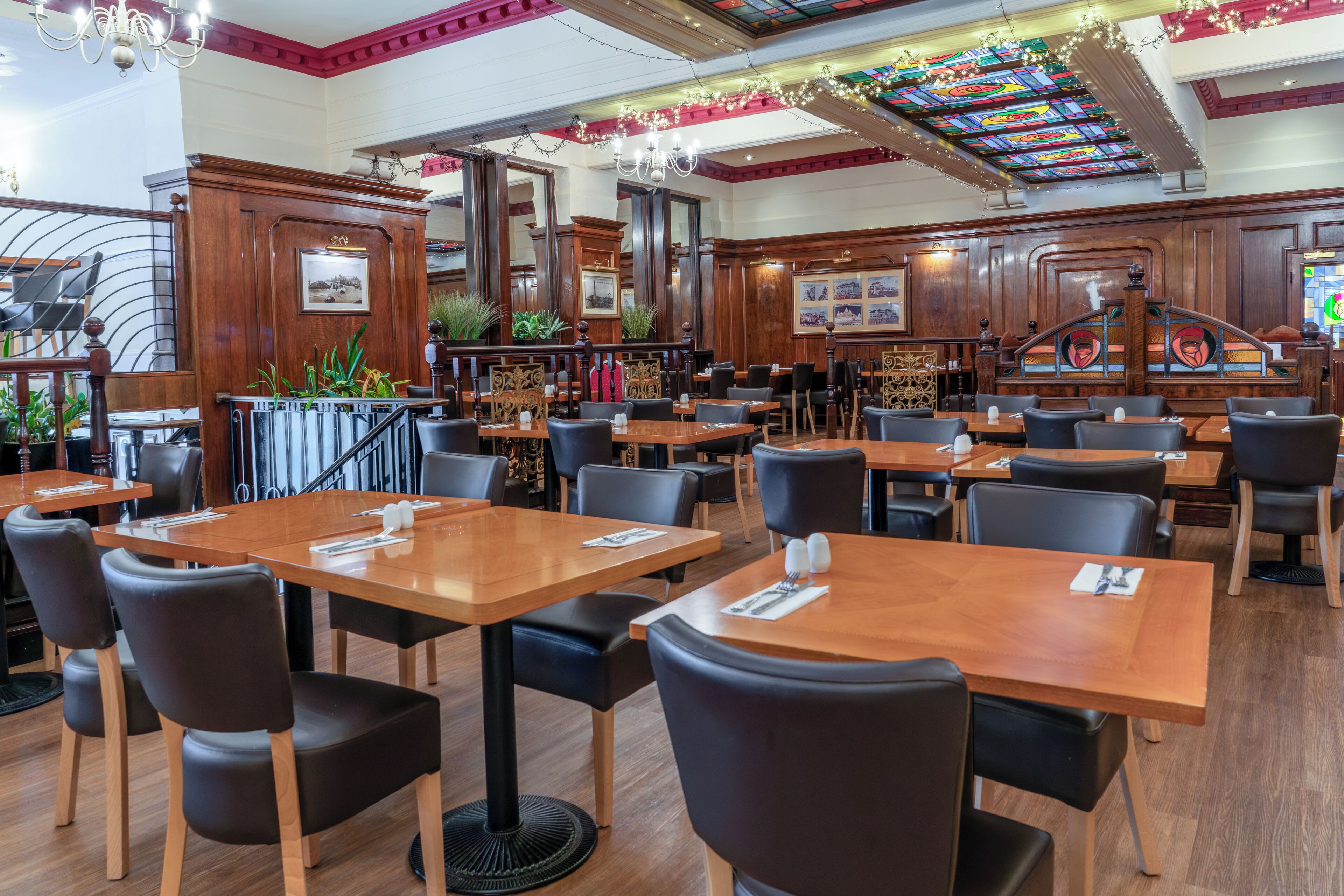 Un restaurante espacioso con mesas de madera y sillas negras, una zona de bar y elementos decorativos como plantas y un techo de vitrales.