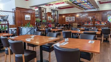 Un restaurante espacioso con mesas de madera y sillas negras, una zona de bar y elementos decorativos como plantas y un techo de vitrales.
