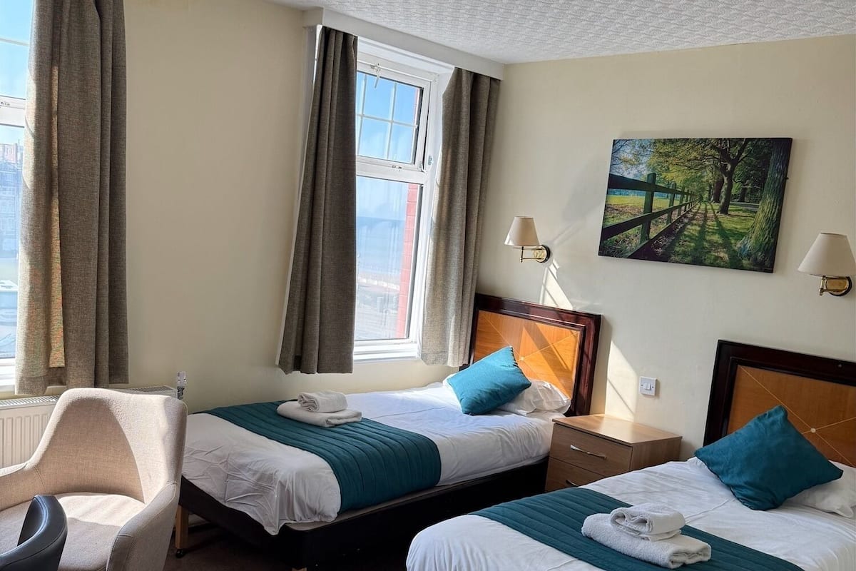 A hotel room with two single beds, a chair, a painting, and a ceiling light fixture.
