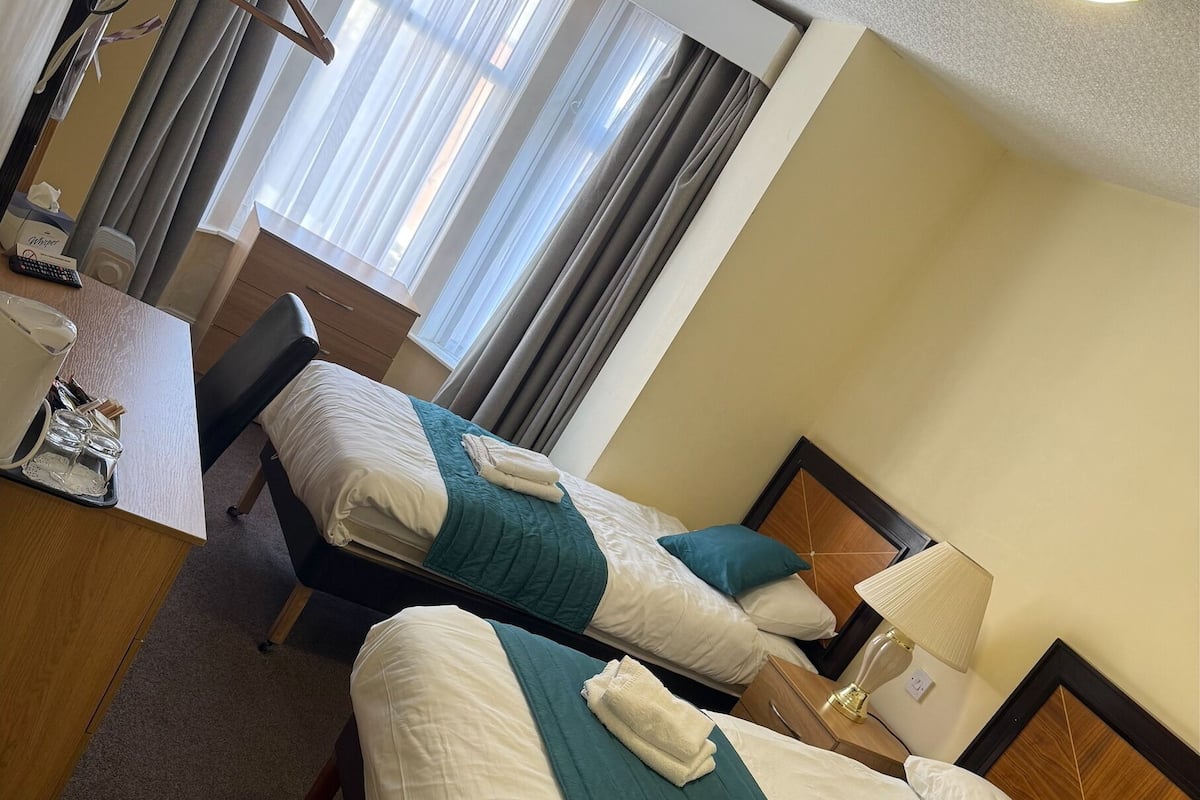 A hotel room with two beds, each with a teal bedspread and white linens. There is a desk with a lamp, a chair, and a small table with a bottle and glasses. The room has a window with curtains and a ceiling light.
