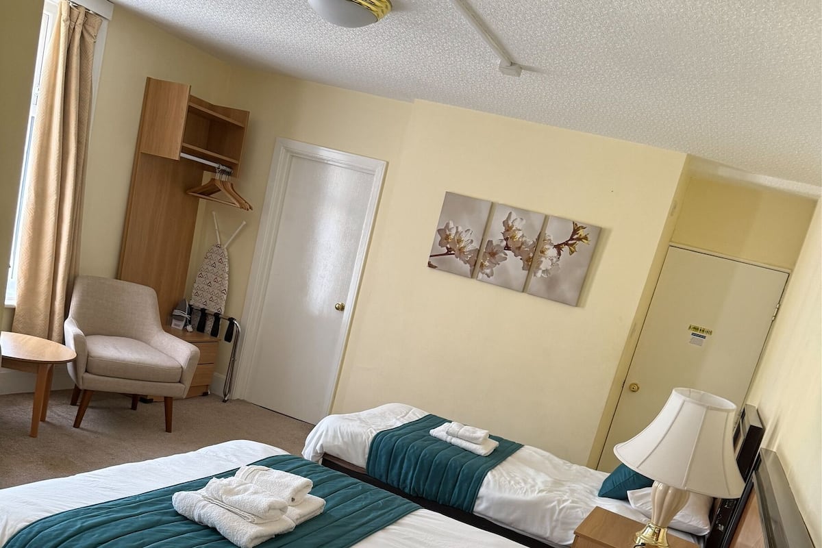 A hotel room with two beds, a chair, a wardrobe, and a ceiling fan.