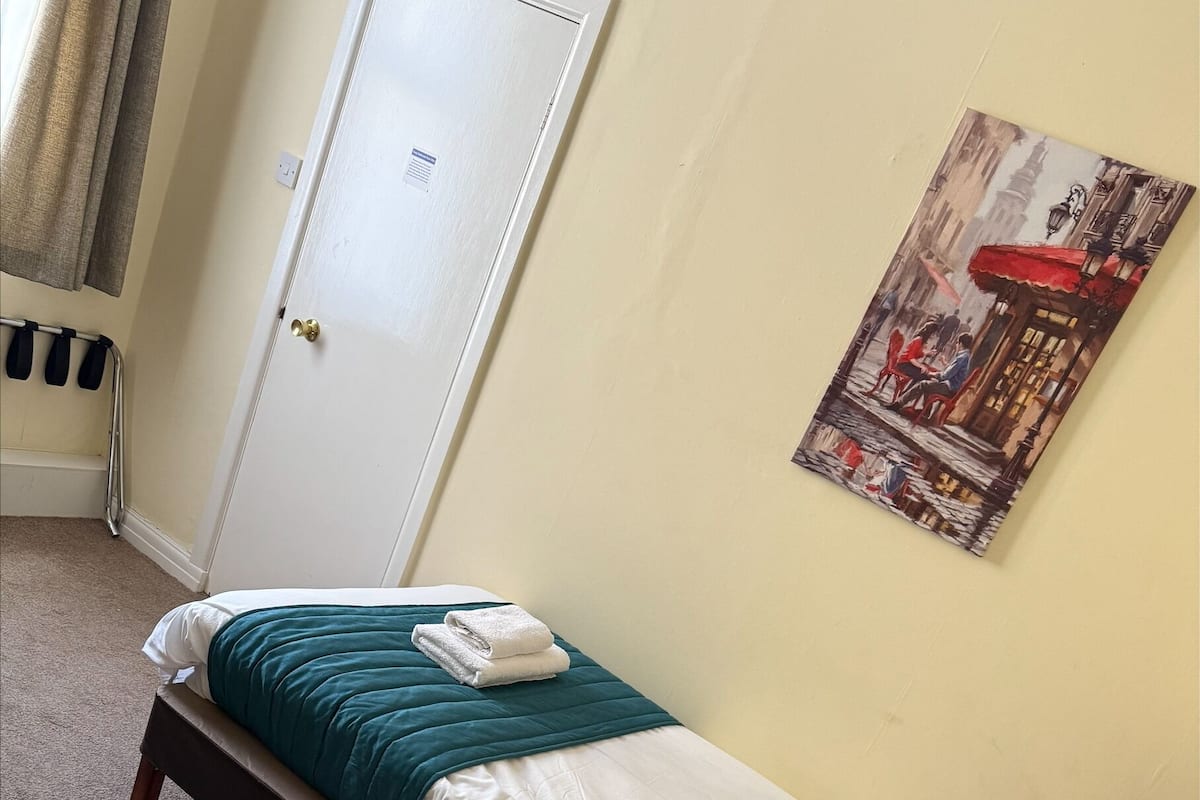 A single bed with a teal blanket and pillows, a wooden nightstand, a door, and a wall-mounted painting.